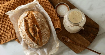 Vers gebakken ambachtelijk brood op bakpapier naast een potje zuurdesemstarter op een houten plank met een lepel