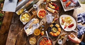 Buitentafel met diverse barbecuegerechten, broodjes, salades, en dranken, met een grill waarop worsten en spiesjes liggen