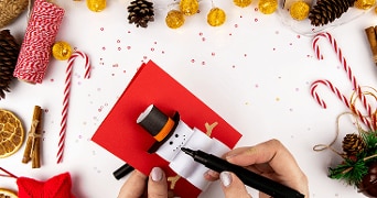 Knutselwerk voor kerst: iemand maakt een sneeuwpop van papier op een rood kaartje, omringd door kerstdecoraties zoals dennentakken, dennenappels, zuurstokken en lichtjes.