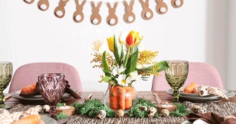 Gedekte paastafel met paarse glazen, borden met croissants, servetten, en decoratie van kunstmos, kwarteleieren en een vaas met voorjaar bloemen en wortels in het midden. Op de achtergrond hangt een slinger met konijntjes aan de muur.