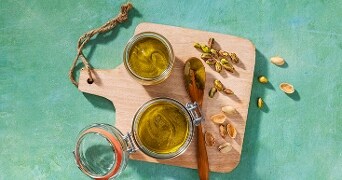 Twee glazen potten met olijfolie en een houten lepel met pistachenoten op een snijplank.