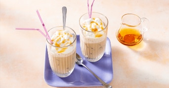 Twee glazen ijskoffie met slagroom en karamelsaus, geserveerd op een paarse schaal met lepels en rietjes. Naast de glazen staat een klein glazen kannetje met karamelsaus.