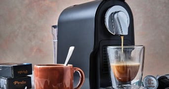 Zwarte koffiemachine met een glas waarin verse koffie wordt gezet, een bruine mok met lepel, en Perla koffiecups op de achtergrond.