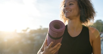 Persoon met een opgerolde paarse fitnessmat in de zon buitenshuis.