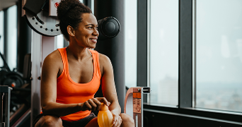 Vrouw in sportkleding met oranje flesje, rustend naast fitnessapparatuur in de sportschool.