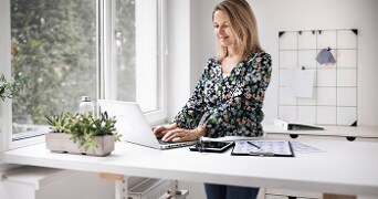 Vrouw werkt op een laptop in een modern kantoor met plantenbak, notitiebord en glazen flesje op het bureau.