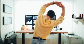 Persoon doet rek oefeningen bij een bureau met computerschermen in een kantooromgeving