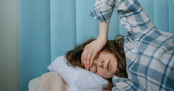 Persoon ligt op bed met pyjama en hand tegen het hoofd, voor een lichtblauw hoofdbord.