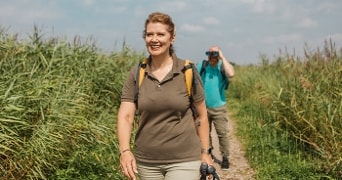 Wandelaars lopen over een pad tussen hoge groene grassen op een zonnige dag, met één persoon die een verrekijker vasthoudt.