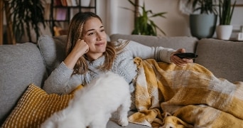 Persoon zit ontspannen op een bank met een afstandsbediening, onder een geel geruite deken, met een witte kat op schoot