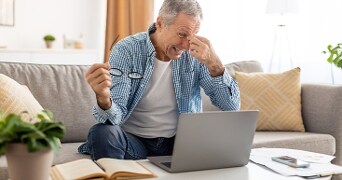 Man zit op een bank met een laptop op schoot, houdt een bril in de hand en leest een open boek, met papieren en een smartphone op tafel.