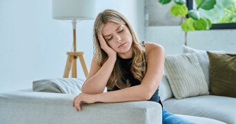 Vrouw zittend op een moderne bank in een lichte woonkamer met sierkussens en een vloerlamp op de achtergrondâ