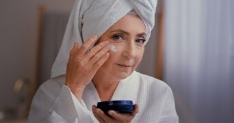 Persoon in badjas en handdoek om hoofd brengt crème aan op gezicht