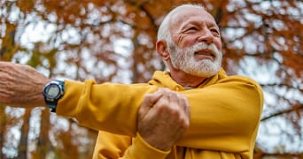 Oudere man in gele trui doet rekoefeningen buiten in het park met herfstbladeren op de achtergrond