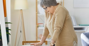 Oudere vrouw leunt voorover op een tafel in een huiselijke woonkameromgeving