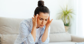 Vrouw in een gestreept overhemd zit op een bank in een lichte woonkamer met een plant op de achtergrond.