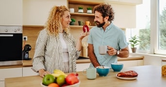 Ontbijtscène in een moderne keuken met een vrouw die een rode appel vasthoudt en een man met een mok, vers fruit en ontbijtproducten op het aanrecht