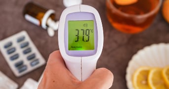 Hand houdt een digitale thermometer vast die een lichaamstemperatuur van 37°C aangeeft, met op de achtergrond medicatie, een glas thee en een schijfje citroen.