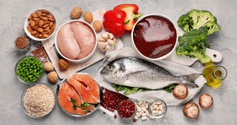 Verschillende gezonde voedingsmiddelen, waaronder kipfilet, rode paprika, broccoli, noten, zalm, dorade, lever, erwten, champignons, bonen, havermout, knoflook en olijfolie, uitgestald op een grijze ondergrond