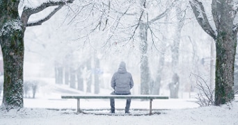 Persoon met winterjas zit op een bankje in een besneeuwd park, omringd door bomen met sneeuw.