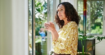 Vrouw in geel wit patroon shirt kijkt uit het raam en houdt een mok vast in een zonnige kamer met planten