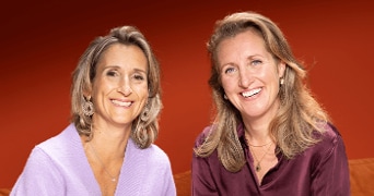 Twee vrouwen in kleurrijke kleding voor een roodbruine achtergrond.