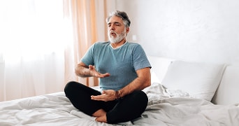 Man in meditatiehouding op bed in een heldere kamer met gordijnen.