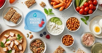 Een assortiment van gezonde snacks zoals magere kwark van Albert Heijn, snackwortels, cherrytomaten, komkommer, gekookte eieren, kipfilet, notenmix, granola repen, walnoten, tonijn, geroosterde kikkererwten, en vers fruit, gepresenteerd op een tafel.