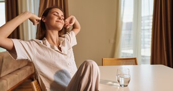 Vrouw in beige kleding zit aan tafel met een glas water, in een woonkamer met natuurlijke lichtinval.