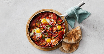 Gerecht met gepocheerde eieren in tomatensaus geserveerd in een pan, vergezeld van broodjes.