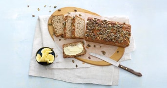 Brood met pompoenpitten op een snijplank, geserveerd met mes en schaaltje boter.