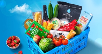 Picknickmand gevuld met producten van Albert Heijn, waaronder AH grillbriketten, 99% energiereep watermeloen, hummus dip, diverse groenten zoals paprika's en komkommer, een kleine watermeloen, en Driscoll's aardbeien, naast andere zomerse producten.