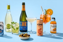Assortiment van alcoholvrije dranken, waaronder een alcoholvrij biertje in glas en blik, een mojito in fles en glas, een fles en glas alcoholvrije bubbelwijn, een spritz cocktail met sinaasappel en kruiden, een glas en flesje spritz, een grapefruit mocktail in blik en een schaaltje groene en zwarte olijven op een lichtblauwe achtergrond.