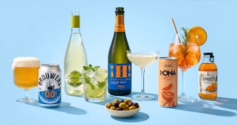 Assortiment van alcoholvrije dranken, waaronder een alcoholvrij biertje in glas en blik, een mojito in fles en glas, een fles en glas alcoholvrije bubbelwijn, een spritz cocktail met sinaasappel en kruiden, een glas en flesje spritz, een grapefruit mocktail in blik en een schaaltje groene en zwarte olijven op een lichtblauwe achtergrond.