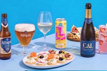 Een feestelijke tafel met verschillende drankjes, waaronder een fles en glas Affligem Blond 0.0 bier, een glas witte wijn, een blikje AH Strawberry Lemon drank, een fles C&L Chloé & Louise Blanc de Blancs mousserende wijn, en een glas roze cocktail met aardbeien en citroen. Op tafel staan ook een bord met luxe hapjes en een schaal met noten en vijgen.