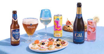 Een feestelijke tafel met verschillende drankjes, waaronder een fles en glas Affligem Blond 0.0 bier, een glas witte wijn, een blikje AH Strawberry Lemon drank, een fles C&L Chloé & Louise Blanc de Blancs mousserende wijn, en een glas roze cocktail met aardbeien en citroen. Op tafel staan ook een bord met luxe hapjes en een schaal met noten en vijgen.