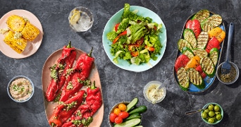 Een kleurrijke presentatie van gegrilde en rauwe groenten, waaronder gevulde rode paprika's, salade, maïs, courgette en een mix van cherrytomaten en komkommers, geserveerd met dips en olijven.