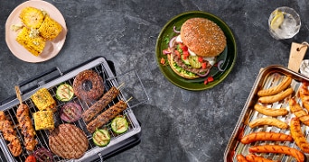 Barbecuegerechten met kipspiezen, hamburgers, groenten en worstjes op een grill, met een bijgerecht van gegrilde maïs en een glas met frisdrank.