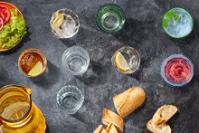 Verschillende glazen met drankjes, een kan, brood, en een bord met salade en tomaat, uitgestald op een zwarte achtergrond.