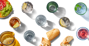 Verschillende glazen met drankjes, een kan, brood, en een bord met salade en tomaat, uitgestald op een zwarte achtergrond.