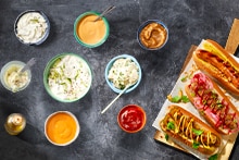 Een afbeelding van verschillende dips en sauzen in schaaltjes, naast drie hotdogs gepresenteerd op een plank, gegarneerd met ui, saus en kruiden.