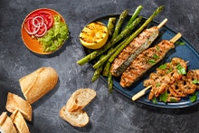 Gegrilde zalmspies, garnalenspies en groene asperges op een blauw bord, geserveerd met gegrilde citroen, salade met tomaat, rode ui en sla, en stukjes stokbrood.