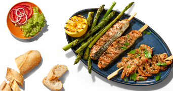 Gegrilde zalmspies, garnalenspies en groene asperges op een blauw bord, geserveerd met gegrilde citroen, salade met tomaat, rode ui en sla, en stukjes stokbrood.