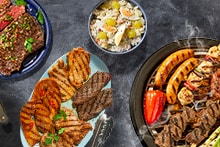 Gegrild vlees op een bord, aardappelsalade in een kom en diverse soorten vlees en groente op een barbecue grill.