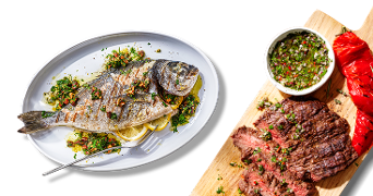 Gegrilde zeebaars op een schaal met kruiden en citroen, naast gegrilde steak met salsa en een rode paprika op een houten plank.