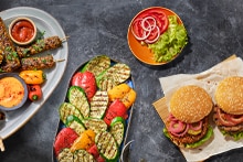 Platte met gegrilde groenten, spiesjes met sausjes, hamburgerbroodjes met sla, tomaat en ui, en een schaaltje met sla, tomaat en ui.