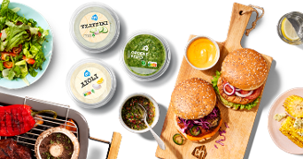 BBQ-omgeving met diverse Albert Heijn producten, waaronder tzatziki, groene pesto, aioli, en op een houten plank gepresenteerde hamburgers. Te zien zijn ook een verse salade, gegrillde biefstuk en champignons, en maiskolf.