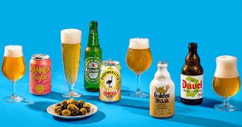Assortiment van verschillende soorten bier, inclusief merken als Heineken, Gulden Draak, Duvel en Mannenliefde, geserveerd in bijpassende glazen met een schaal groene en zwarte olijven op een blauwe achtergrond.