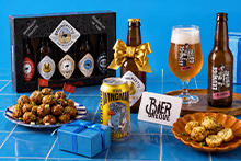 Assortiment met speciaalbieren van Brouwerij ’t IJ en Bird Brewery (Wingman IPA), glas bier, biercadeaupakket, biercheque, borrelhapjes en blauwe cadeauverpakkingen op een blauwe tegeltafel.