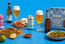 Assortiment van speciaalbieren, waaronder Brand NEIPA, La Trappe Trappist, Uiltje Wizard Wit en Brand Weizen, geserveerd naast borrelhapjes, noedels met groenten en vlees, en een salade, op een blauwe ondergrond.
